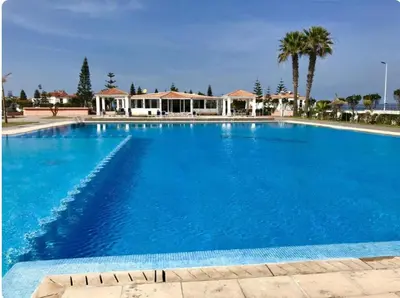 a large blue swimming pool with palm trees and houses at Villa en bord de plage, située dans une résidence à El Mansouria, idéale pour une grande famille ou pour des personnes seules in Mansouria