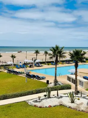 a swimming pool with palm trees and a beach at Sidi Rahal Front mer, piscine & accès direct plage in Dar Oulad Hajj Kassem