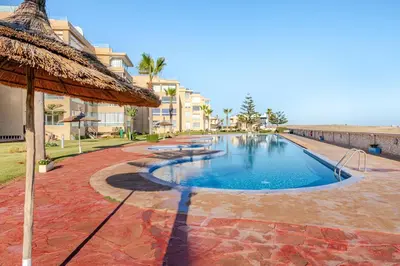 a swimming pool with a straw umbrella next to a building at Appartement de Luxe - Piscine et Accès à la Plage in Bouznika