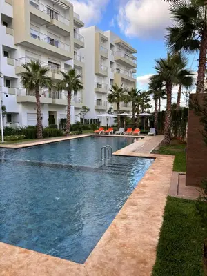 a swimming pool in front of a building with palm trees at Appartement Bouznika POOL & BEACH in Bouznika