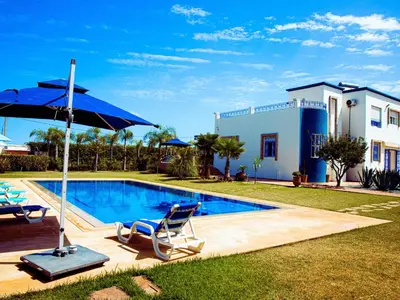 a swimming pool with a blue umbrella and chairs and a house at Instant Saisons - Ferme d'hôtes in Skhirat